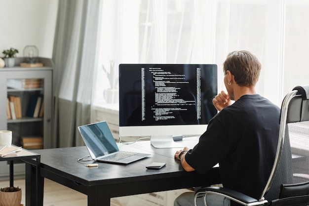 Rear view of computer programmer sitting at the table in front of computer monitor and developing software at office
