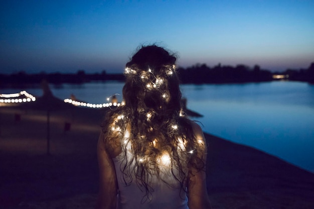 rear view of woman with illuminated string lights standing at beach against sky