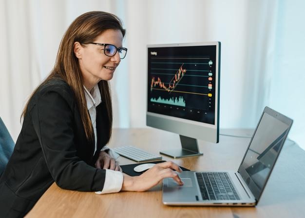 Side view of cheerful female broker trading online on binary option while sitting at table and browsing netbook