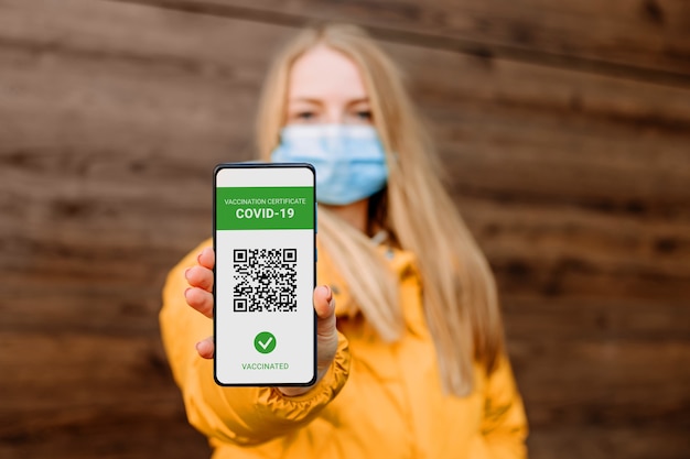 Studentka kavkazského původu v lékařské masce ukazuje obrazovku mobilního telefonu s aplikací o očkování proti koronaviru, covid-19, dřevěné pozadí