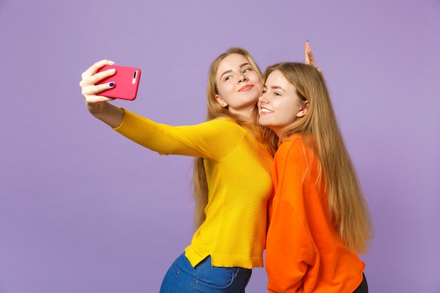 Two smiling young blonde twins sisters girls in colorful clothes doing selfie shot on mobile phone isolated on pastel violet blue wall . People family lifestyle concept. 