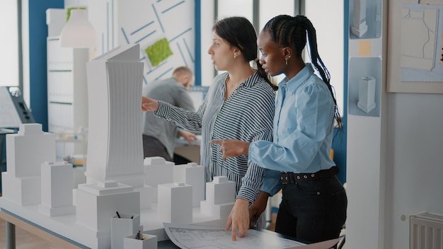 Workmates analyzing blueprints plan and building model on table, working on construction. Women creating architectural structure to design project development with layout sketch.