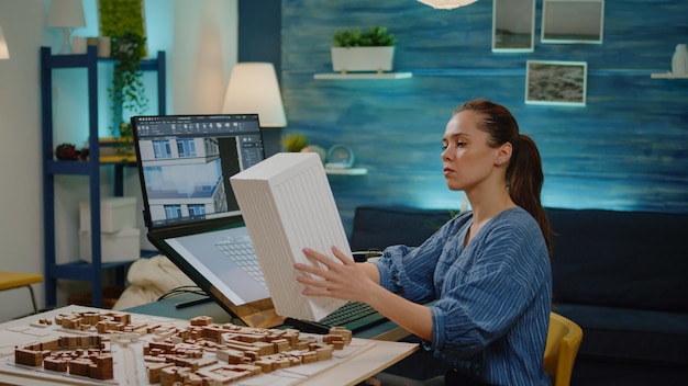 young architect holding maquette model in hand working on building layout for construction development. engineer woman with blueprint plan analyzing touch screen monitor in office.