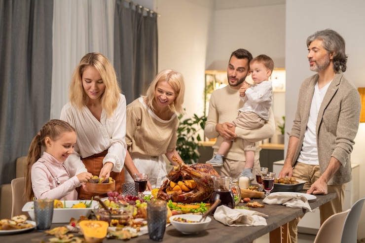 Fotografie young mother giving her cute little daughter fresh fruits while both standing by served festive table on background of the rest of family