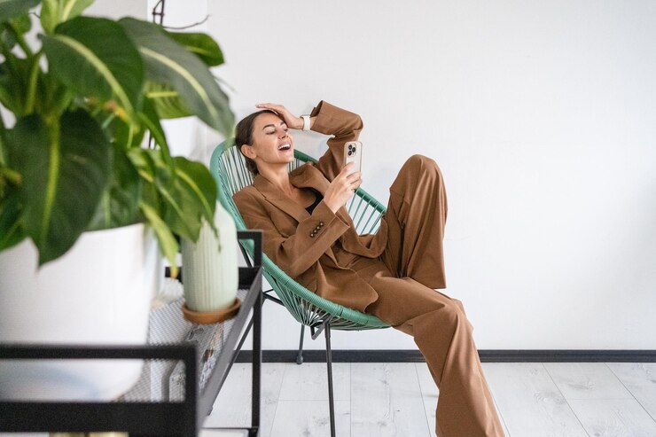 Fotografie young woman using phone and laughing at camera she is wearing stylish suit and seating in chair