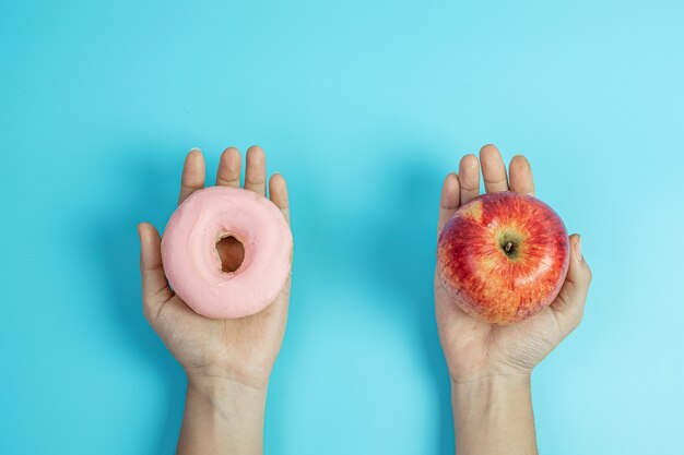 Žena drží červené jablko a růžový donut, žena si vybírá mezi ovocem, které je zdravé, a sladkostmi, což jsou nezdravé potraviny. Koncept diety, obezity, stravovacích návyků a výživy