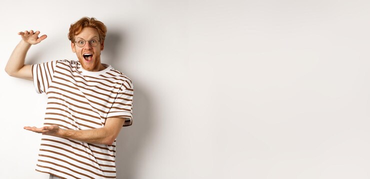 Zdarma Fotografie amazed redhead man in glasses showing something large size and smiling at camera saying wow standing