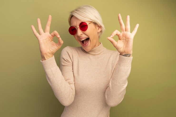 Zdarma Fotografie confident young blonde woman wearing sunglasses looking at front doing ok sign isolated on olive green wall