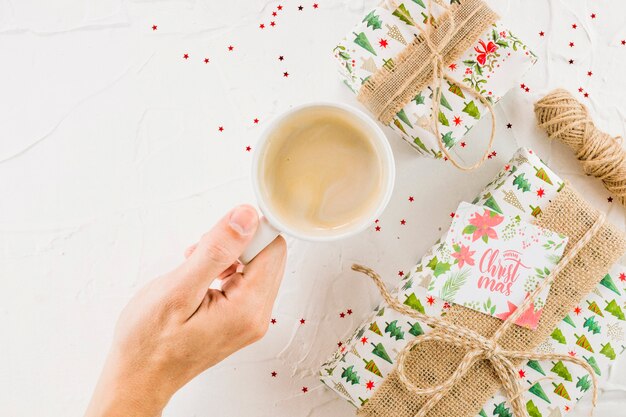 Hand with cup near present boxes and post card with Christmas inscription