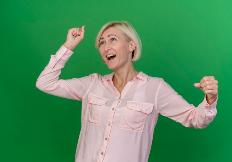 Zdarma Fotografie joyful young blonde slavic woman looking up and clenching fists isolated on green background
