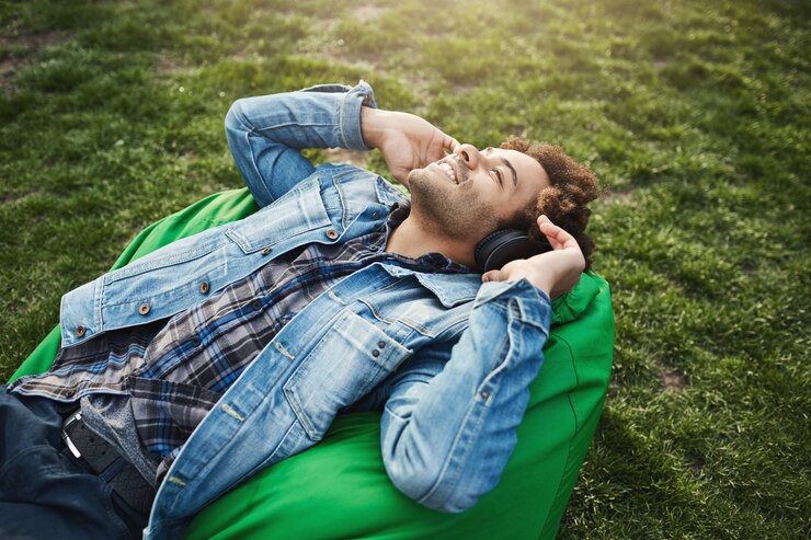 Zdarma Fotografie side-view portrait of happy attractive african-american with afro hairstyle lying on bean bag chair while listening music and looking at sky with relaxed and pleased expression, spending time in park