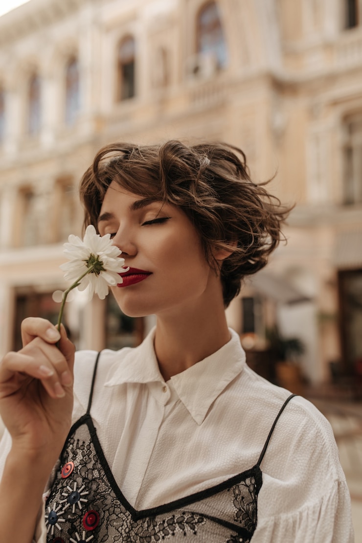 Zdarma Fotografie wavy-haired woman in light shirt with black lace sniffing flower in city. tender woman with red lips and short hair poses at street.