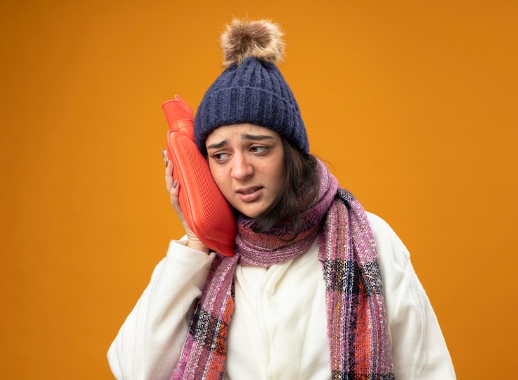 Zdarma Fotografie weak young caucasian ill girl wearing robe winter hat and scarf touching head with hot water bag looking at side isolated on orange background with copy space