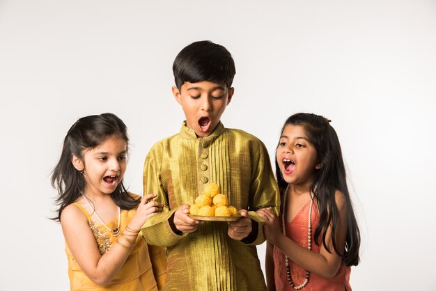 3 crianças ou irmãos indianos em trajes tradicionais sentados no sofá ou no fundo branco, segurando presentes e doces ou laddu