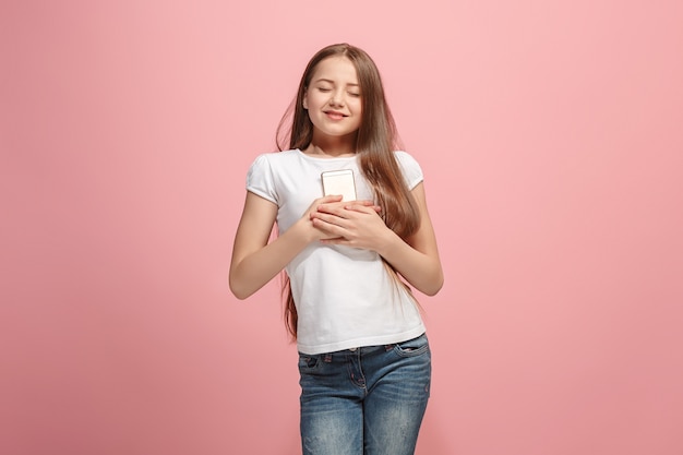 A adolescente feliz com o telefone em pé e sorrindo contra a parede rosa