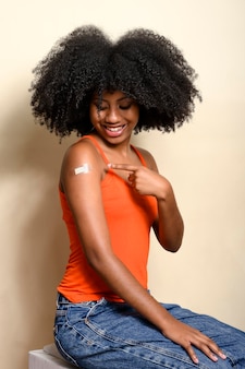 A adolescente negra aponta para o adesivo em seu braço, sorri e mostra que foi vacinada, isolada em fundo bege.