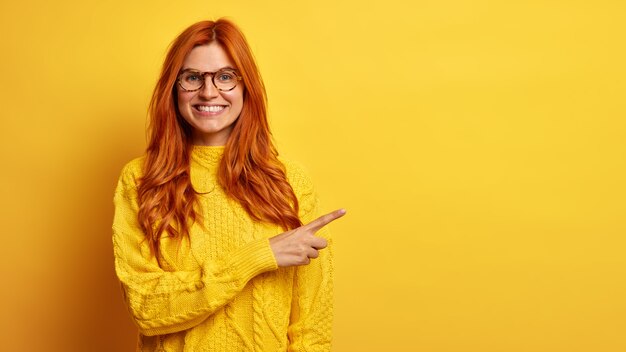 A bela mulher ruiva positiva sorri amplamente aponta para o espaço em branco e recomenda uma boa oferta de venda vestida com uma camisola de malha.