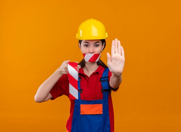 A boca da jovem construtora confiante selada com fita de advertência segura a fita e mostra o gesto de parar em um fundo laranja isolado com espaço de cópia