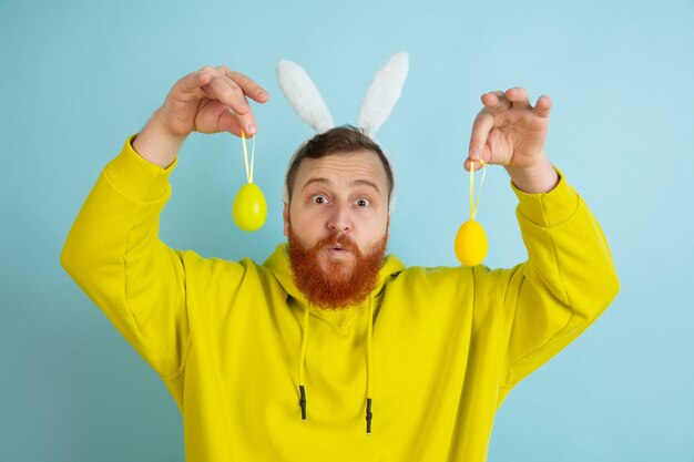 A caça ao ovo está chegando. Homem caucasiano como um coelhinho da Páscoa com roupas casuais brilhantes sobre fundo azul do estúdio. Saudações de Páscoa feliz. Conceito de emoções humanas, expressão facial, feriados. Copyspace.