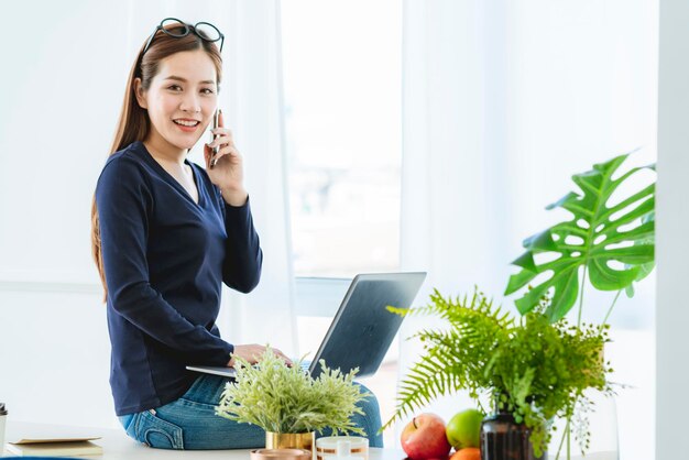 A camiseta preta da mulher de negócios asiáticos casuais criativos trabalha em casa com smartphone e laptop com luz matinal da janela