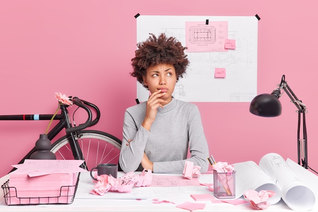 A cena horizontal de uma mulher afro-americana pensativa em um espaço de coworking tem uma expressão pensativa que faz o projeto arquitetônico desenhar esboços para projetar um novo edifício, faz planejamentos ou suposições