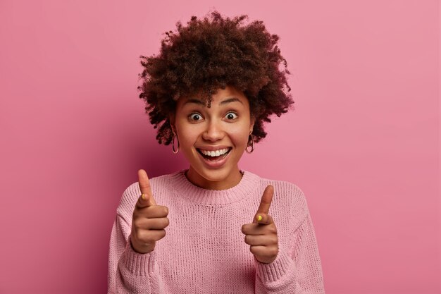 A cena horizontal de uma mulher muito alegre aponta diretamente, sugere se juntar a ela, escolhe você, sorri amplamente, vestida com um macacão casual, posa sobre uma parede rosada. Pessoas e conceito de escolha