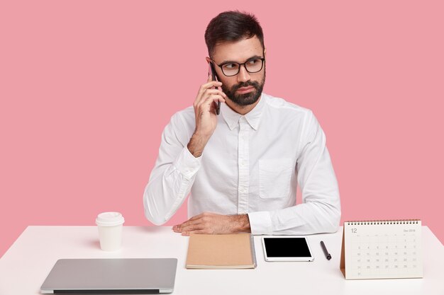 A cena horizontal do empresário sério tem cerdas grossas, conversa telefônica, foco à distância, vestido com roupas formais, tem pedido na mesa