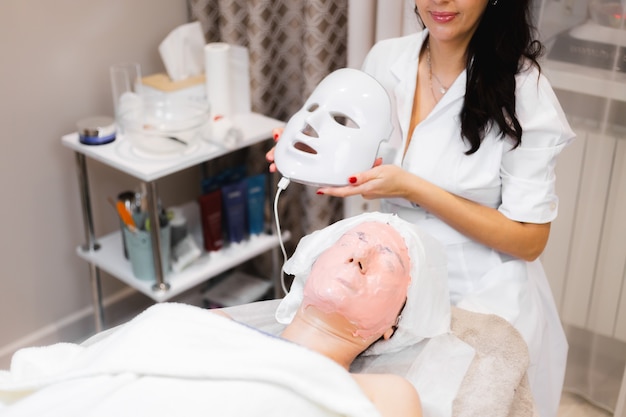 A cliente encontra-se no salão, na mesa de cosmetologia, com uma máscara branca no rosto