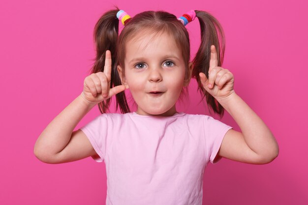 A criança emocional surpresa e impressionada coloca os dedos para cima, abre a boca com espanto, olhando atentamente. Pouco modelo engraçado posa vestindo casual luz rosa camiseta, scrunchies coloridos.