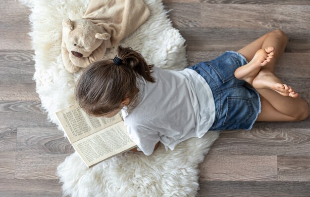 A criança lê um livro deitado sobre um tapete aconchegante em casa com seu ursinho de pelúcia de brinquedo favorito.