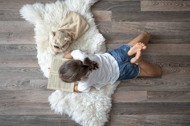 A criança lê um livro deitado sobre um tapete aconchegante em casa com seu ursinho de pelúcia de brinquedo favorito.