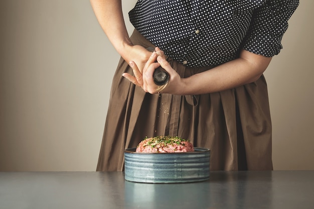 A dona de casa adiciona um pouco de pimenta na carne picada em uma tigela de cerâmica na mesa azul