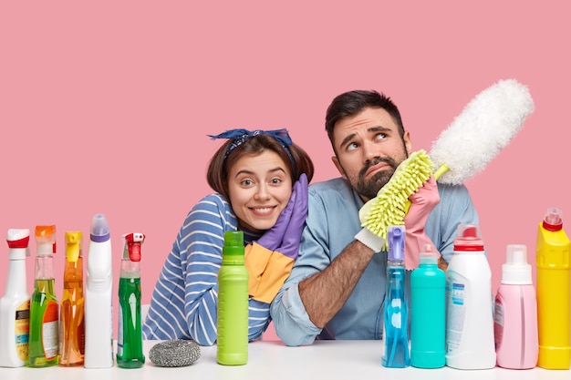 A dona de casa feliz se inclina sobre o ombro do marido atencioso, limpa a casa juntos, faz a limpeza, cercada de detergentes de limpeza, segura o esfregão, sala de poeira tem pouco descanso após um trabalho cansativo.