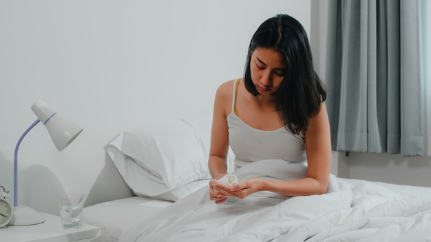 A fêmea indiana doente e insalubre sofre de insônia. Jovem asiática tomando medicamento analgésico para aliviar a dor de cabeça e beber um copo de água, sentada na cama em seu quarto em casa de manhã.