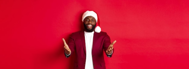 Foto grátis a festa de natal e o conceito de férias feliz homem negro com chapéu de papai noel lhe dão as boas-vindas apontando as mãos para