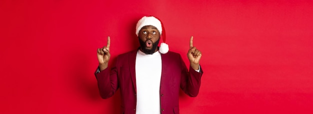 A festa de natal e o conceito de férias impressionaram o homem negro com chapéu de papai noel apontando os dedos para cima dizendo uau