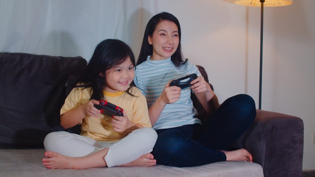 A filha e a família asiáticas jovens jogam jogos em casa na noite. Mãe coreana com menina usando momento feliz engraçado joystick juntos no sofá na sala de estar. Mãe engraçada e criança adorável estão se divertindo