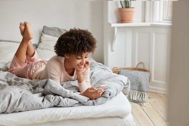 A foto de uma mulher relaxada de pele escura assiste a história online no celular, lê a postagem com informações interessantes, vestida com roupas casuais casuais, deita-se na cama, tem uma expressão feliz. Interior aconchegante