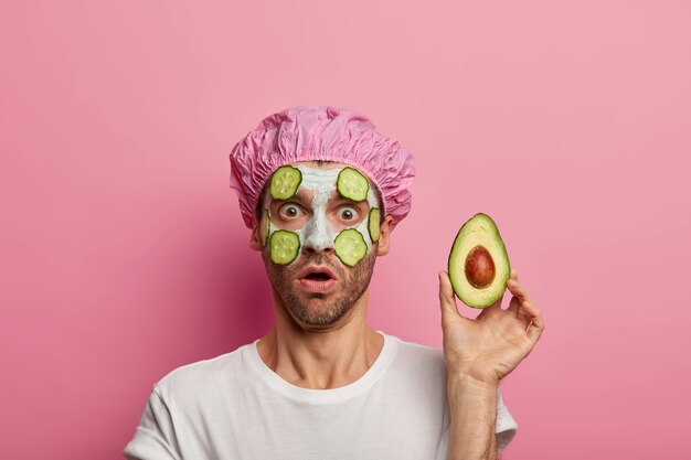 A foto do homem atordoado engasga de admiração, segura o abacate perto do rosto, surpreendido pelo efeito da máscara de argila, usa touca de banho, tem cerdas, isolado sobre a parede rosa do estúdio. Beleza, bem-estar