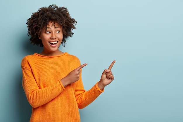 A foto horizontal agradou a mulher de pele escura com corte de cabelo afro, aponta para longe com os dois dedos da frente, mostra um espaço em branco para sua promoção, isolado sobre a parede azul. Pessoas, conceito de propaganda