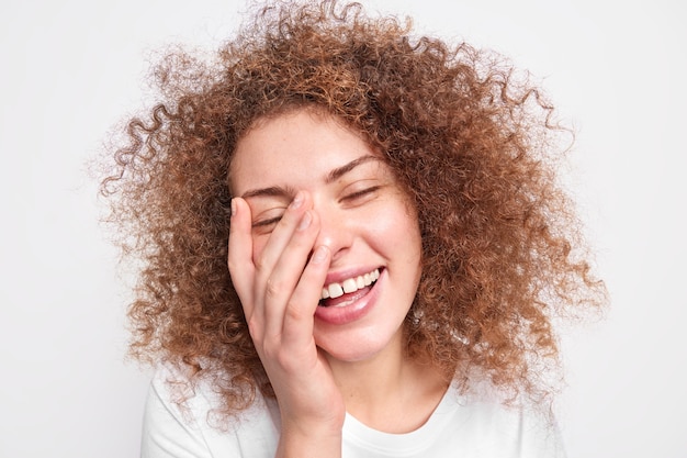 A foto na cabeça de uma linda mulher europeia sincera com cabelo crespo e encaracolado se diverte, ri, cobre o rosto com as palmas das mãos, sorri amplamente expressa felicidade isolada sobre uma parede branca. Emoções positivas