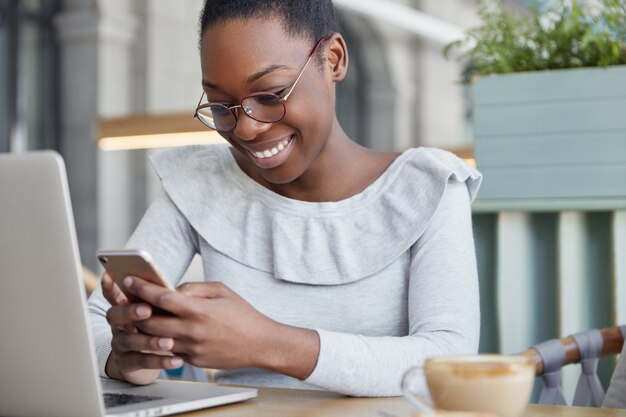 A foto recortada de um redator feminino satisfeito lê informações positivas no telefone inteligente, senta-se em frente a um laptop aberto e bebe café aromático.