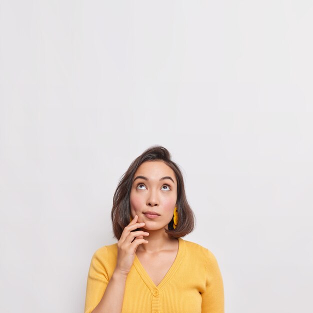 A foto vertical de uma jovem asiática pensativa e sonhadora com cabelo escuro focado acima considera que algo usa um macacão amarelo casual isolado sobre o espaço de cópia de parede branca para seu anúncio