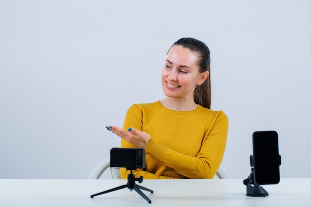 A garota blogueira sorridente está apontando para a esquerda com a mão sentada na frente de sua mini câmera no fundo branco