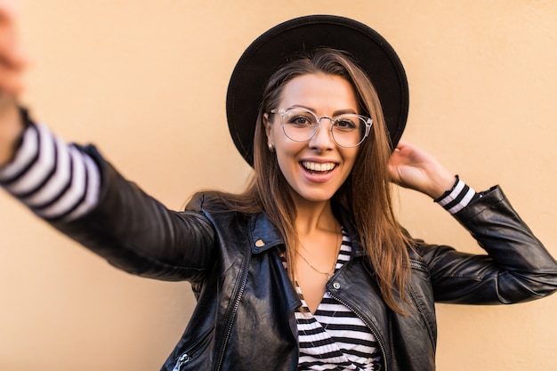 A garota bonita da moda em jaqueta de couro e chapéu preto faz selfie isolada na parede amarela clara