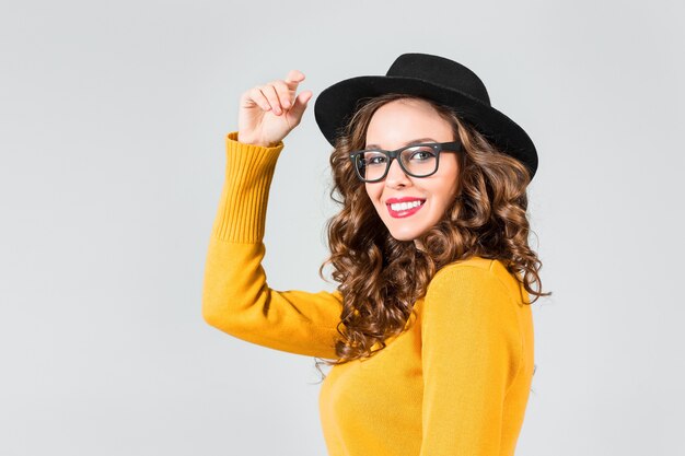 A garota feliz de óculos e chapéu na parede cinza do estúdio