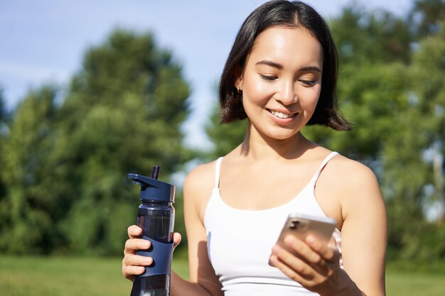 A garota fitness sorridente bebe água, verifica seu aplicativo no smartphone e parece feliz, fica hidratada em fogo