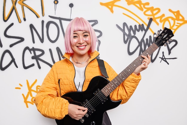 A guitarrista alegre faz poses de cabelo rosa com um instrumento musical e usa uma jaqueta laranja