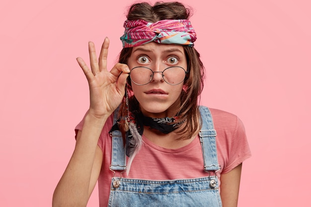 A hippie surpreendida usa bandana e macacão, olha escrupulosamente através dos óculos, lê algo incomum, fica encostada na parede rosa. mulher hippie emocionada e entorpecida em pé