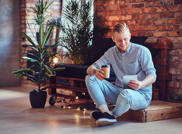 A imagem de corpo inteiro de um homem elegante loiro vestido com uma camisa de lã e jeans usando um tablet PC e bebe café em uma sala de estar.